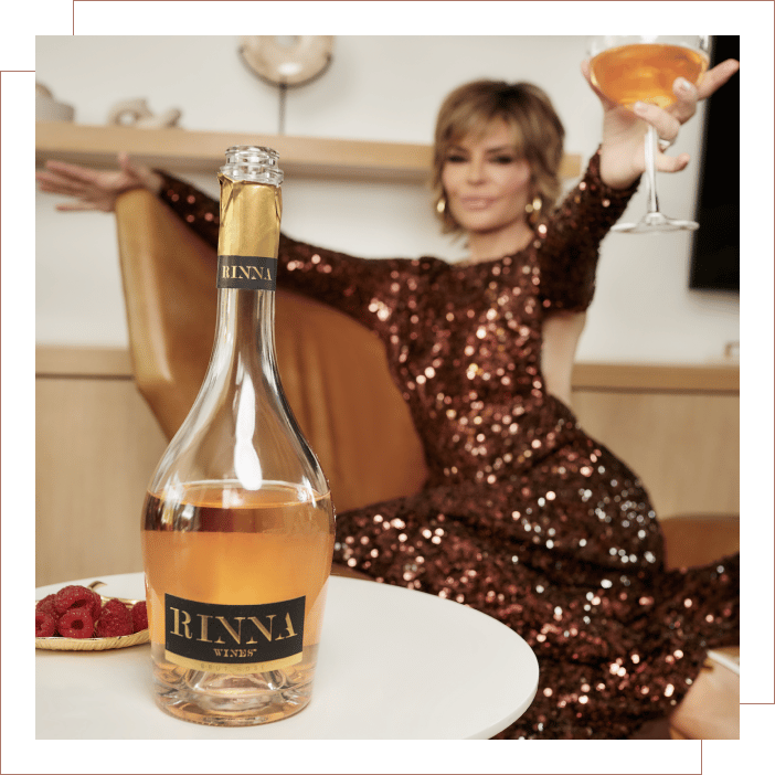 A bottle of Rinna Wines Brut open in the foreground on a table with Lisa Rinna in the background raising a glass.