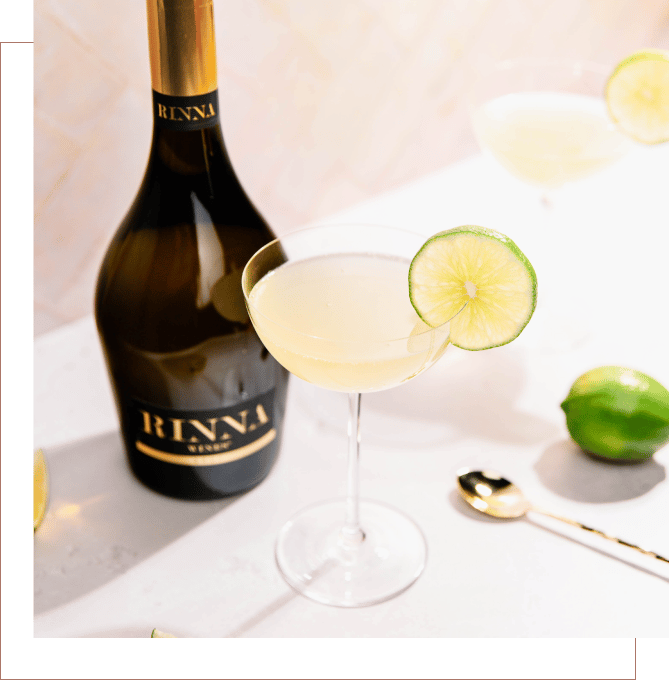 A Rinna margarita in a class with a lime wedge garnish next to a bottle of Rinna Wine Brut sparkling wine